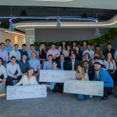 A group of approximately 25 young adults are posing for a photo in an office setting. Some are holding large novelty checks with various amounts written on them. The individuals are diverse in appearance and are dressed in business casual attire.  Artificial greenery lines the wall. The mood of the image is celebratory and suggests a competition or award ceremony.
