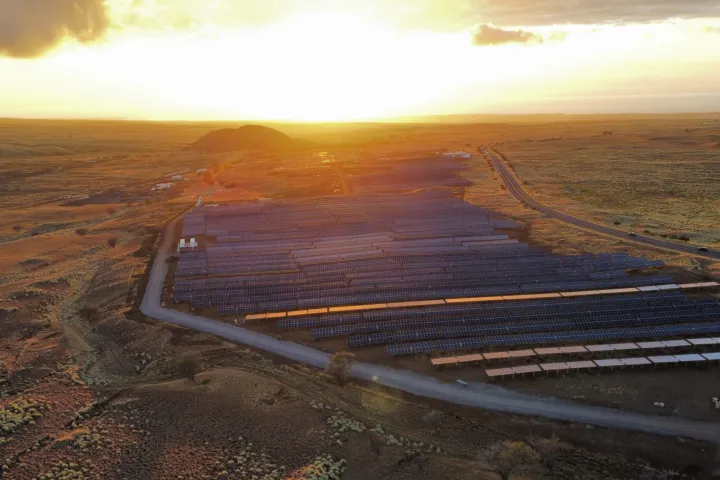 Aerial view of a large solar farm in a vast open landscape at sunset, with rows of solar panels capturing sunlight and a road running alongside the facility.