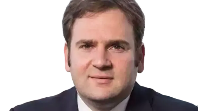 Man in a dark business suit with a blue patterned tie, sitting indoors with a blurred cityscape visible through the window behind him.