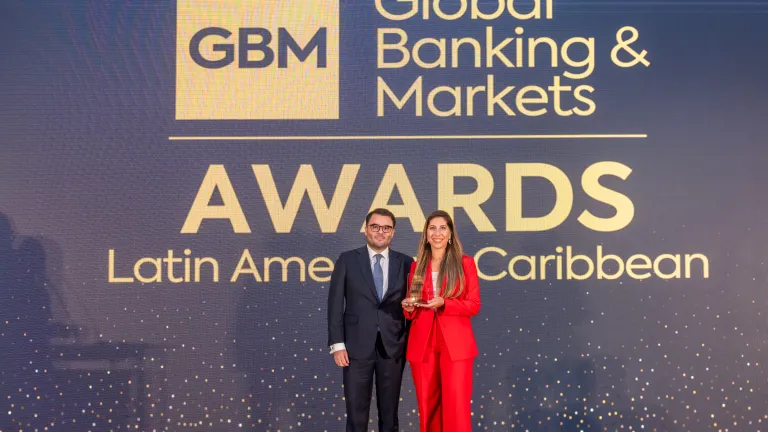 A man in a suit and a woman in a red suit stand on stage holding an award at the Global Banking & Markets Awards for Latin America and the Caribbean, with a large event backdrop behind them.