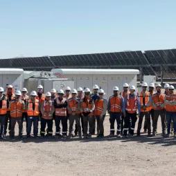 Group of workers in orange safety vests and white helmets standing in front of solar panels and equipment at a solar energy facility under clear skies.