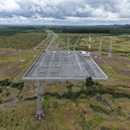 Aerial view of an electrical substation surrounded by green fields and transmission towers, with a cloudy sky and distant forests.