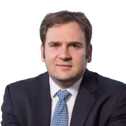 Man in a dark business suit with a blue patterned tie, sitting indoors with a blurred cityscape visible through the window behind him.