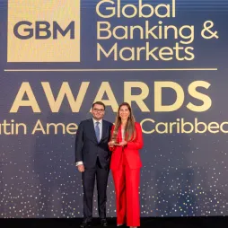 A man in a suit and a woman in a red suit stand on stage holding an award at the Global Banking & Markets Awards for Latin America and the Caribbean, with a large event backdrop behind them.